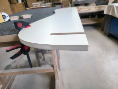 Bir zanaatkar atölyenin içinde düzgün kıvrımlı bir tezgah şekillendirir. Aletler ve ahşap malzemeler düzenlenerek, özel mobilya yapımında kullanılan hassasiyet ve beceriyi sergiliyorlar..