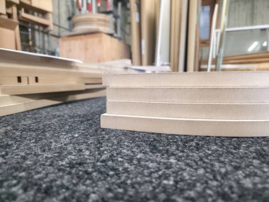 Bir atölyede düzgünce yığılmış çeşitli odun parçaları var. Döşeme halı kaplı ve araçlar arka planda görünüyor. Bu da etkin bir inşaat ortamını gösteriyor..