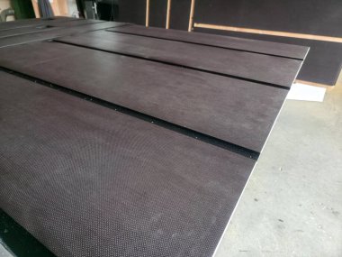 Koyu, desenli yüzey panelleri atölye masasına düzgün bir şekilde dizilmiş. Ayarlar, yaklaşan bir inşaat projesine hazırlık olduğunu gösteriyor. Arkaplanda araçlar ve malzemeler görünür.