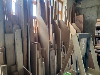 Çeşitli ahşap tahtalar ve tahtalar düzgün bir şekilde düzenlenir ve iyi aydınlatılmış bir atölyede istiflenir. Güneş ışığı pencerelerden süzülerek inşaat malzemeleriyle dolu alanı aydınlatıyor..