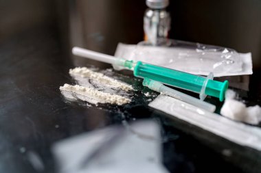 Sıvı ve toz maddelerle dolu şırıngalar yansıtıcı bir yüzeye yerleştirilir. Çevre loş bir şekilde aydınlanmış. Bu da geceleri gizli bir aktivitenin gerçekleştiğini gösteriyor..