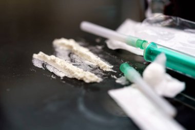 Sıvı ve toz maddelerle dolu şırıngalar yansıtıcı bir yüzeye yerleştirilir. Çevre loş bir şekilde aydınlanmış. Bu da geceleri gizli bir aktivitenin gerçekleştiğini gösteriyor..