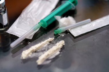 Sıvı ve toz maddelerle dolu şırıngalar yansıtıcı bir yüzeye yerleştirilir. Çevre loş bir şekilde aydınlanmış. Bu da geceleri gizli bir aktivitenin gerçekleştiğini gösteriyor..