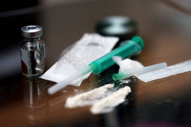 Sıvı ve toz maddelerle dolu şırıngalar yansıtıcı bir yüzeye yerleştirilir. Çevre loş bir şekilde aydınlanmış. Bu da geceleri gizli bir aktivitenin gerçekleştiğini gösteriyor..