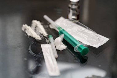 Sıvı ve toz maddelerle dolu şırıngalar yansıtıcı bir yüzeye yerleştirilir. Çevre loş bir şekilde aydınlanmış. Bu da geceleri gizli bir aktivitenin gerçekleştiğini gösteriyor..