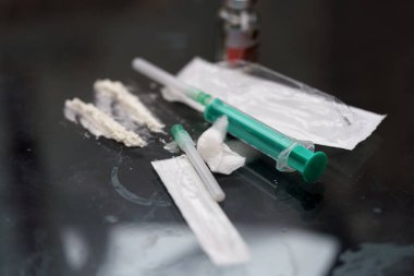 Sıvı ve toz maddelerle dolu şırıngalar yansıtıcı bir yüzeye yerleştirilir. Çevre loş bir şekilde aydınlanmış. Bu da geceleri gizli bir aktivitenin gerçekleştiğini gösteriyor..