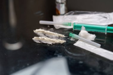 Sıvı ve toz maddelerle dolu şırıngalar yansıtıcı bir yüzeye yerleştirilir. Çevre loş bir şekilde aydınlanmış. Bu da geceleri gizli bir aktivitenin gerçekleştiğini gösteriyor..