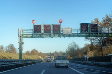 Elektronik işaretler hız limitlerini gösterirken araçlar öğleden sonra yoğun bir otoyolda yol alırlar. Bölge ağaçlar ve açık mavi gökyüzüyle çevrili. Güvenli sürüş koşullarını sağlıyorlar..