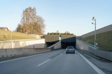 Bir araba yeşil ve yumuşak bir yamaçla çevrili karanlık bir tünel girişine yaklaşıyor. Parlak gökyüzü, ilerideki gölgeli açıklıkla çelişiyor. Bu da kentsel bir ortamı gösteriyor..