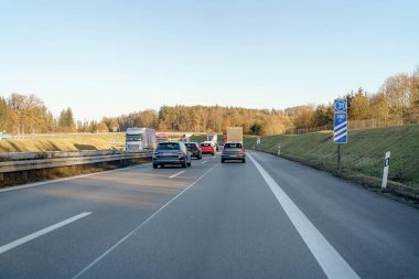 Araçlar, açık bir gökyüzünün altındaki yeşil ağaçlar tarafından çerçevelenmiş çok şeritli bir otoyol boyunca sürüyorlar. Sahne, yol yolculuğunun huzurlu öğleden sonra atmosferini yakalıyor..