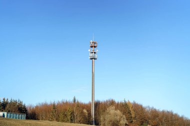Parlak mavi bir gökyüzünün altındaki seyrek ağaçlarla çevrili kırsal bir alanda bir iletişim kulesi belirgin bir şekilde yükseliyor. Sahne, modern bağlantıyı yansıtan sakin ve açık bir atmosfer yakalıyor..