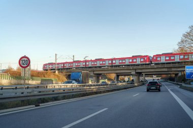 Alacakaranlıkta yoğun bir otoyolun üzerindeki yüksek bir köprüden geçen kırmızı bir tren. Araçlar güneş batarken yol boyunca hareket ediyor, manzaraya sıcak bir ışık saçıyor..