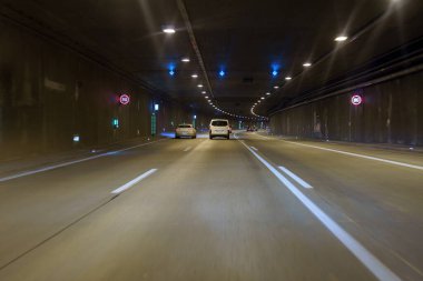 İki araç loş bir tünelde yan yana gidiyor. Mavi ve kırmızı ışıklar duvarları noktalayıp gece yolculuğu için atmosferik bir ortam yaratıyor..