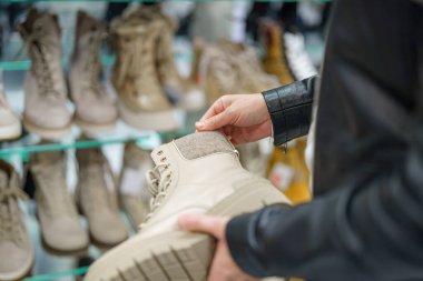 Bir perakende mağazasında, bir birey bir çift yüksek bej botu incelerken bir çift ayakkabı seçimiyle çevrilidir. Boşluk çeşitli biçim ve renklere sahiptir.
