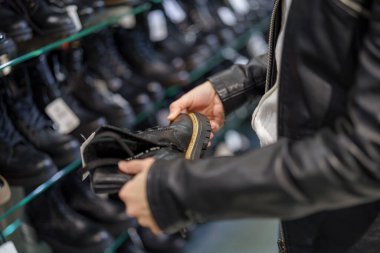 Bir kişi, çeşitli ayakkabı stilleriyle dolu rafları incelerken bir çift siyah botu inceler. Mağaza iyi aydınlatılmış, çizmelerdeki son trendleri sergiliyor..