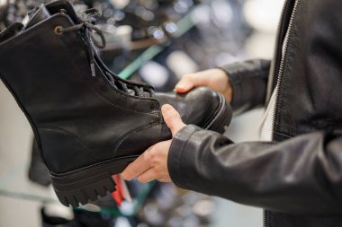 Moda bir mağazada, alışveriş yapan kişi siyah bir botu dikkatle inceler, deri bir tasarım ve diğer ayakkabı seçenekleriyle çevriliyken engebeli bir tabanı gösterir..