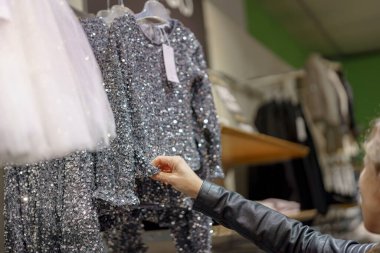 Bir kişi, sergilenen çeşitli şık kıyafetlerle çevrili bir butikte parıldayan, pullu bir çocuk kıyafetini inceliyor. Titrek kumaş ışığı yansıtıyor ve göz kamaştırıcı bir etki yaratıyor..