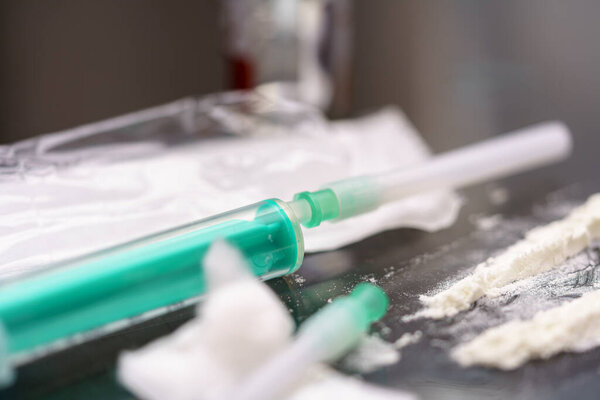 A used syringe rests on a surface alongside white powdery substances, indicating drug use in a somber, dimly lit environment that highlights addiction struggles.