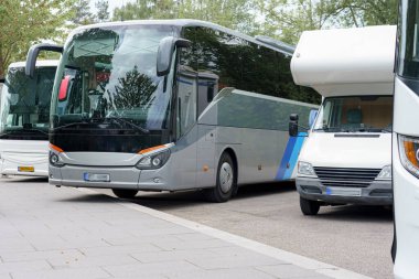 Bir belediye otobüsü sakin bir park yerinde bir karavanın yanına park edilmiş. Güneş parlıyor, araçları aydınlatıyor ve çevresindeki yeşillikleri aydınlatıyor..