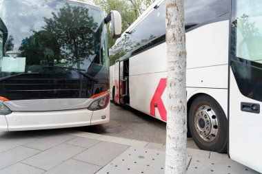 Gündüz vakti yoğun bir otobüs terminalinde yan yana duran iki büyük otobüs. Bölgede yeşillik ve modern kentsel ortam yer alıyor..