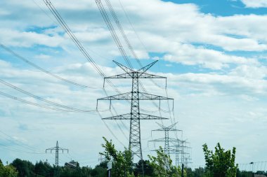 Yeşil kırsal bir arazide, açık mavi gökyüzünün altındaki uzak tepeleri birbirine bağlayan yüksek elektrik hatları yükseliyor. Sahne modern altyapının ortasındaki huzurlu bir anı yakalıyor..