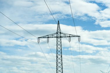 Uzun bir iletim kulesi tüylü bulutların ve mavi gökyüzünün zeminine çarpıyor ve kırsal kesimde elektrik dağıtımı için altyapıyı vurguluyor..