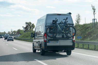 Arkasına güvenli bir şekilde bisiklet monte edilmiş açık bir otoyolda giden bir karavan. Kabarık bulutlar açık mavi gökyüzünü doldururken ağaçlar yol kenarına dizilmiş, yolculuk için hoş bir günü işaret ediyor..