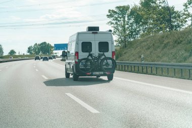 Beyaz bir minibüs açık gökyüzünün altında bir otoyol boyunca gidiyor, arka rafta iki bisiklet taşıyor. Çevresindeki araçlar yeşilliklerle çevrili olarak yolda ilerliyorlar..