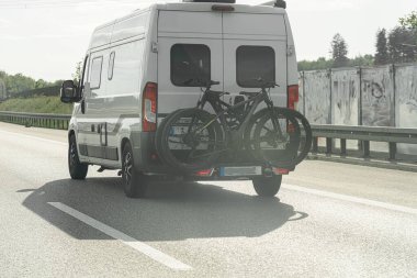 Beyaz bir minibüs otoyol boyunca gidiyor, arkasında iki bisiklet taşıyor. Öğleden sonra güneşi, çevreleyen yeşil araziye sıcak bir ışık saçıyor..