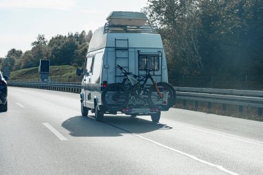 Beyaz bir karavan, açık gökyüzünün altındaki bir otoyolda ilerliyor. Arka taraftaki askıda bisikletleri taşıyor. Manzarada yolu kaplayan ağaçlar var..