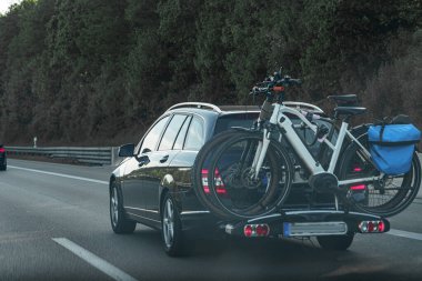 İki bisiklet taşıyan bir bisiklet parkı ile donatılmış bir otoyolda seyahat eden bir araba görülüyor. Çevrede öğleden sonra saatlerinde berrak bir gökyüzünün altında yemyeşil ağaçlar bulunur..