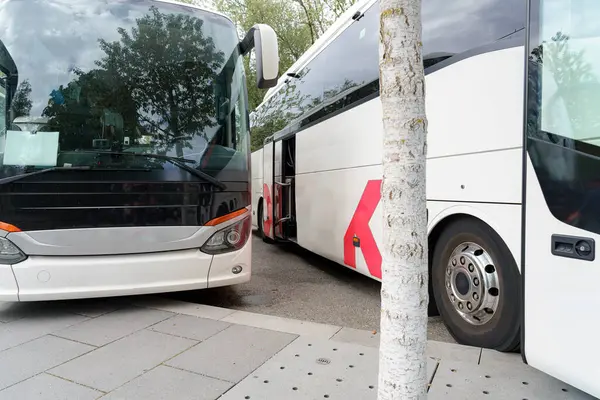 Gündüz vakti yoğun bir otobüs terminalinde yan yana duran iki büyük otobüs. Bölgede yeşillik ve modern kentsel ortam yer alıyor..