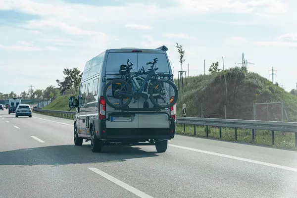 Arka rafa monte edilmiş bisikletlerle donatılmış bir minibüs, açık mavi bir gökyüzünün altında otoyol boyunca ilerliyor. Bereketli yeşil manzara, keyifli yolculuk atmosferine katkı sağlar..