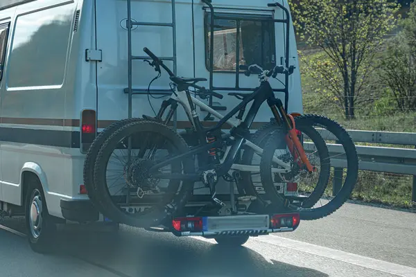 İki dağ bisikleti, yeşil manzaralarla çevrili güneşli bir otoyol boyunca beyaz bir minibüsün arkasına bağlanmış bir askıda güvenle dururlar..