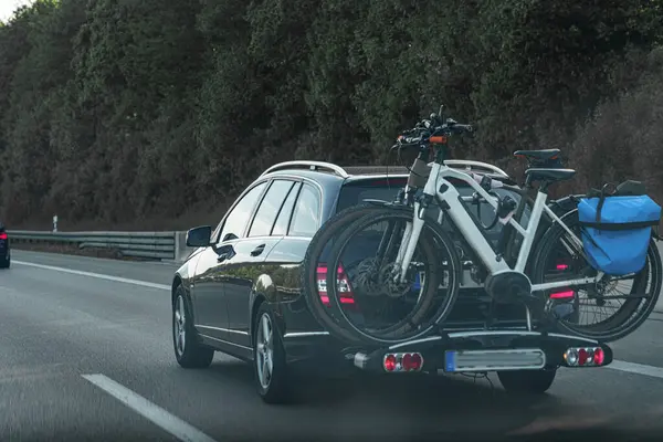 İki bisiklet taşıyan bir bisiklet parkı ile donatılmış bir otoyolda seyahat eden bir araba görülüyor. Çevrede öğleden sonra saatlerinde berrak bir gökyüzünün altında yemyeşil ağaçlar bulunur..