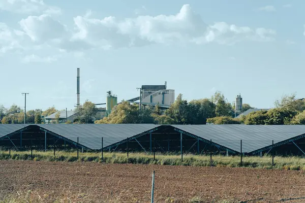 Güneş panelleri kahverengi bir alan boyunca uzanırken, büyük bir endüstriyel tesis bulutlarla dolu mavi bir gökyüzüne karşı duruyor. Mekan huzurlu ama endüstriyel..