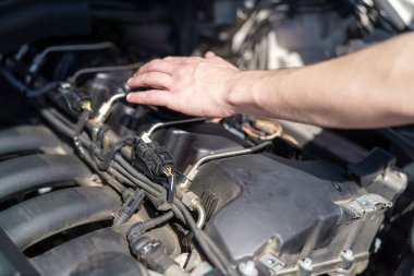 Bir tamirci bir aracın motorunu inceler, çeşitli bileşenleri ve kabloları kontrol eder. Parlak gün ışığı çalışma alanını aydınlatır, detaylara dikkat çeker.