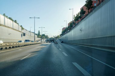 Araçlar berrak mavi gökyüzünün altındaki modern bir şehir tünelinde sorunsuzca seyahat ederler. Çevresindeki yeşil alanlar ve binalar gün boyunca rahatlatıcı bir atmosfer yaratıyor..