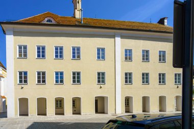 Tarihi bir binada uzun pencereler ve açık mavi bir gökyüzünün altında gün ortası güneşini yansıtan sıcak renkli bir cephe bulunur. Şehir ortamında klasik mimari sergileniyor.