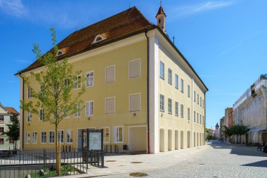 Öğlen vakti açık mavi bir gökyüzünün altında ağaçlar ve dükkanlarla çevrili tarihi bir bina, kaldırım taşı bir caddede tek başına duruyor. Mimari, büyüleyici ve klasik bir tasarım sergiliyor..