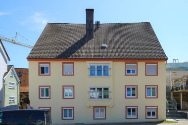 Üç katlı bir konut binasında güneş ışığını yansıtan sarı ve beyaz renklerden oluşan bir karışım sergileniyor. Yakınlarda bir inşaat alanı bölgede devam eden gelişmeleri gösteriyor..