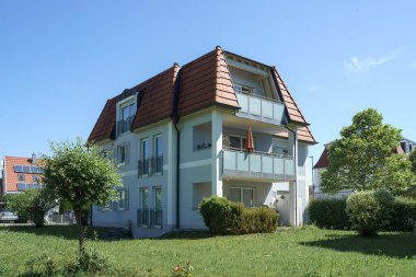 Çağdaş, üç katlı, eğimli çatılı bir bina yemyeşil ve mavi gökyüzünün ortasında duruyor. Mülkte balkonlar ve bakımlı peyzajlar yer alıyor, aile yaşamı için ideal..