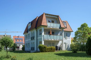 Güneşli bir mahallede, yemyeşil çimenler ve ağaçlarla dolu modern bir yerleşim yeri bulunmaktadır. Mimaride balkonlar ve parlak mavi gökyüzünün altında eğimli bir çatı yer alıyor..