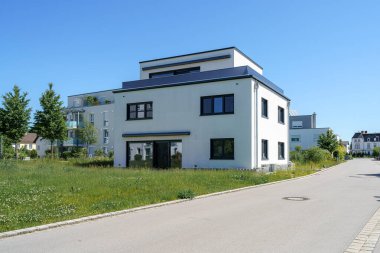 Çağdaş bir yerleşim yeri, çiçek açan kır çiçekleri ve çimlerle çerçevelenmiş sakin bir caddede duruyor. Açık gökyüzü, gündüz atmosferine mükemmel bir dokunuş ekler..