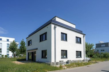 Bereketli yeşilliklerin arasında, geniş pencereler ve açık mavi gökyüzünün altında minimalist estetik bir yapı bulunur. Çevre huzurlu ve davetkar..