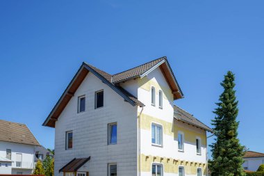 Kısmen yenilenmiş dış görünüşü olan bir yerleşim yeri parlak mavi bir gökyüzüne karşı duruyor. Çevrede yeşil alanlar ve diğer evler var. Huzurlu bir mahalle ortamı yaratıyorlar..