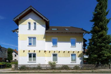 Banliyö mahallesindeki bir binada, dış yüzeyi sarı yalıtım malzemeleriyle kaplanırken yenileme izleri görülüyor. Parlak mavi gökyüzü neşe dolu atmosfere katar.