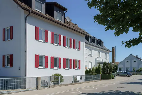 İki yerleşim yeri, beyaz cephelere karşı kırmızı ve yeşil panjurları olan sessiz bir cadde boyunca uzanır. Parlak gökyüzü huzurlu atmosferi vurguluyor.