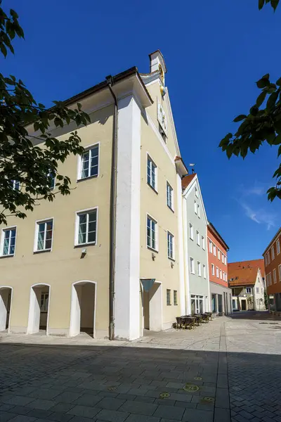 Güneş, renkli cepheli ve açık havada oturmaya davet edilen büyüleyici binaların yer aldığı pitoresk bir kasaba meydanının üzerinde ışıl ışıl parlıyor. Ağaçlar seyircilere gölge sağlar..