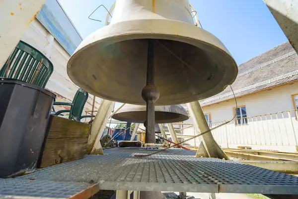 Metal ızgara ve kırsal bir çevreyle çevrili iki büyük çan ön planda belirgin şekilde durur. Güneş ışığı, yıpranmış yüzeylerini vurguluyor. Bu sessiz yerin cazibesini ortaya çıkarıyor..